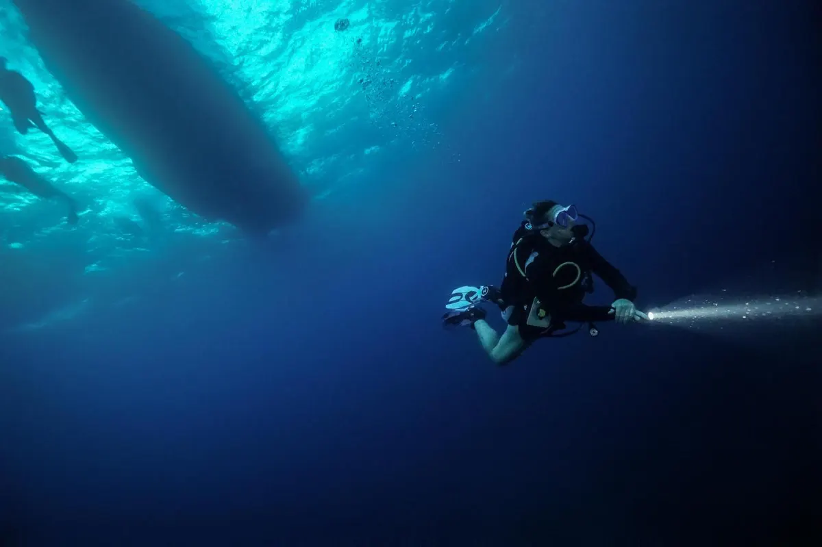 Diver descending with a torch beam beneath the boat as the light fades