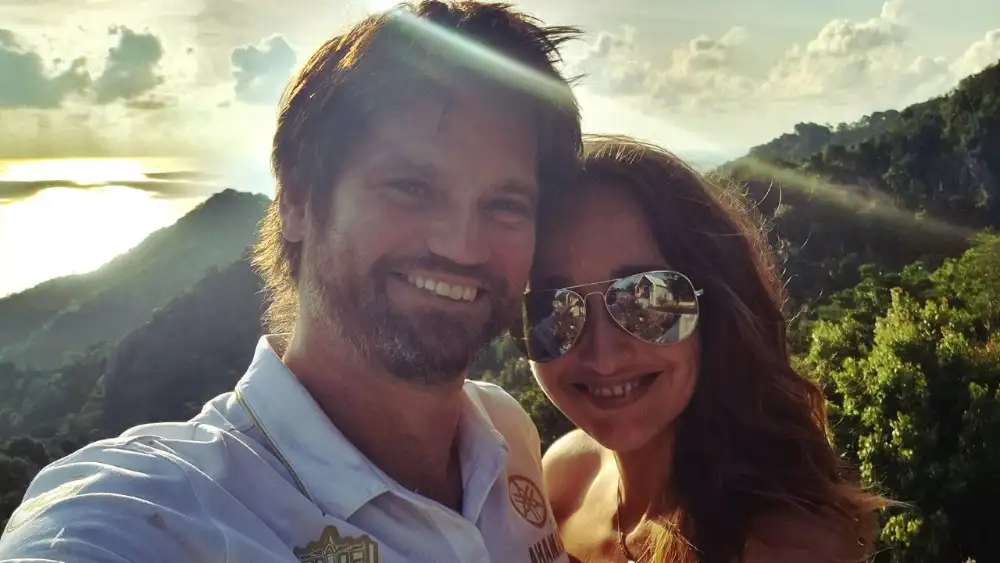 A smiling couple taking a selfie at a hilltop viewpoint on Koh Tao at sunset, with jungle-covered hills and the sea glowing behind them