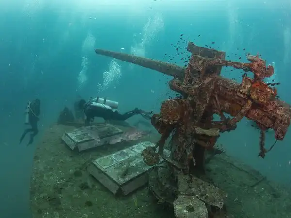 Two divers swimming past the coral-encrusted deck gun of the HTMS Sattakut wreck, Koh Tao