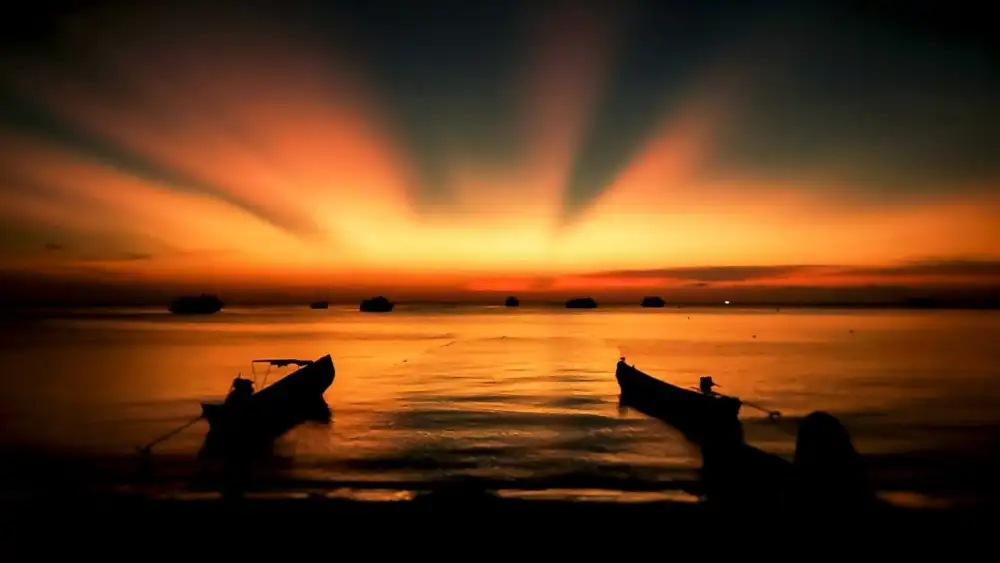 Two longtail boats silhouetted against a fiery orange sunset on a Koh Tao beach, with dive boats on the horizon
