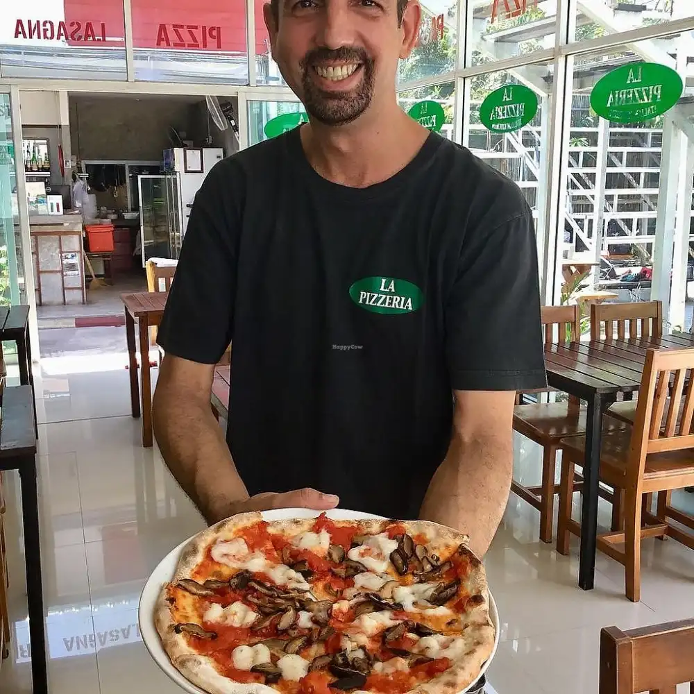 The owner of La Pizzeria da Claudio smiling and holding a freshly baked mushroom pizza in the restaurant's dining room