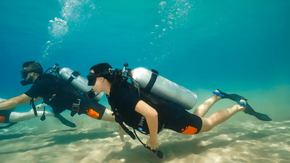 Two scuba divers in shorty wetsuits swimming over a sandy bottom in shallow turquoise water during training